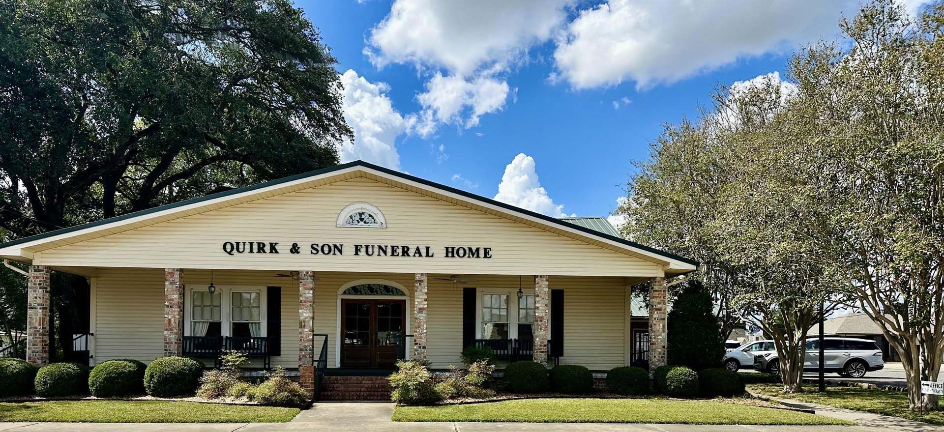 Exterior of Quirk & Son Funeral Home with a beige facade, columns, and a bright blue sky.