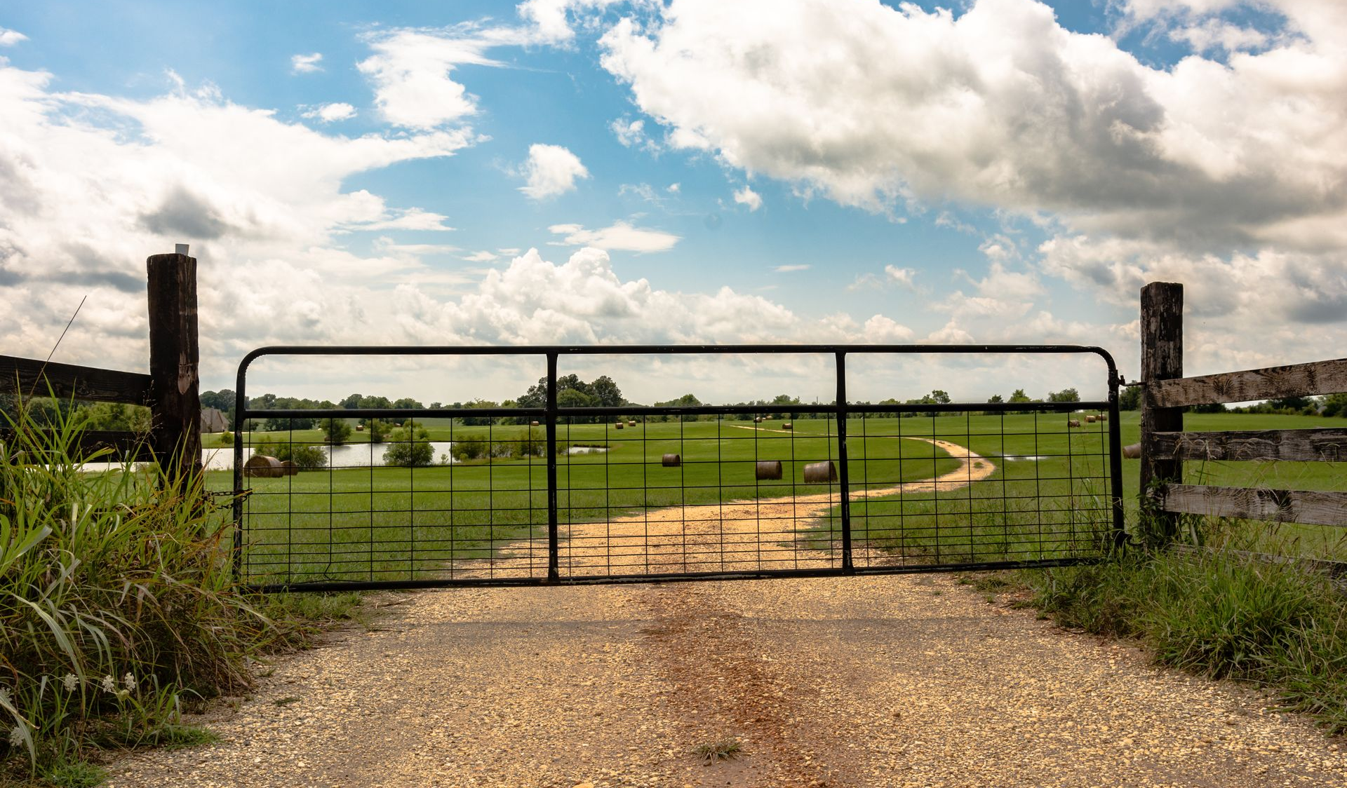 A closed metal gate on a gravel path leading to a grassy field with a pond, under a cloudy sky.