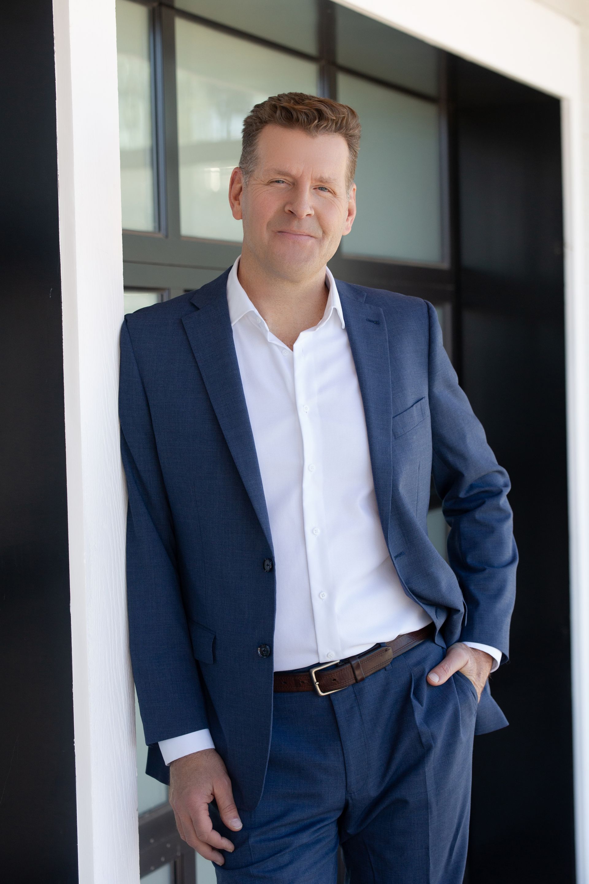 Pete Behrens in a blue suit and white shirt leans against a doorway, looking toward the camera with a neutral expression.