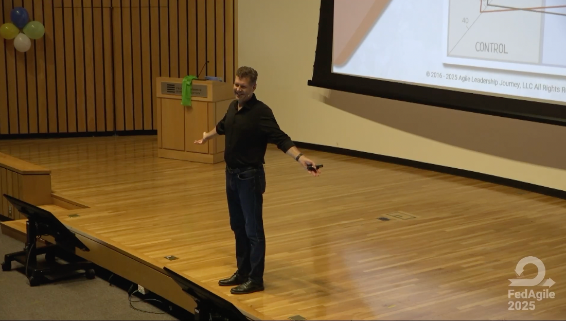 Pete Behrens is delivering keynote in black clothing, standing on a wooden stage at FedAgile 2025.