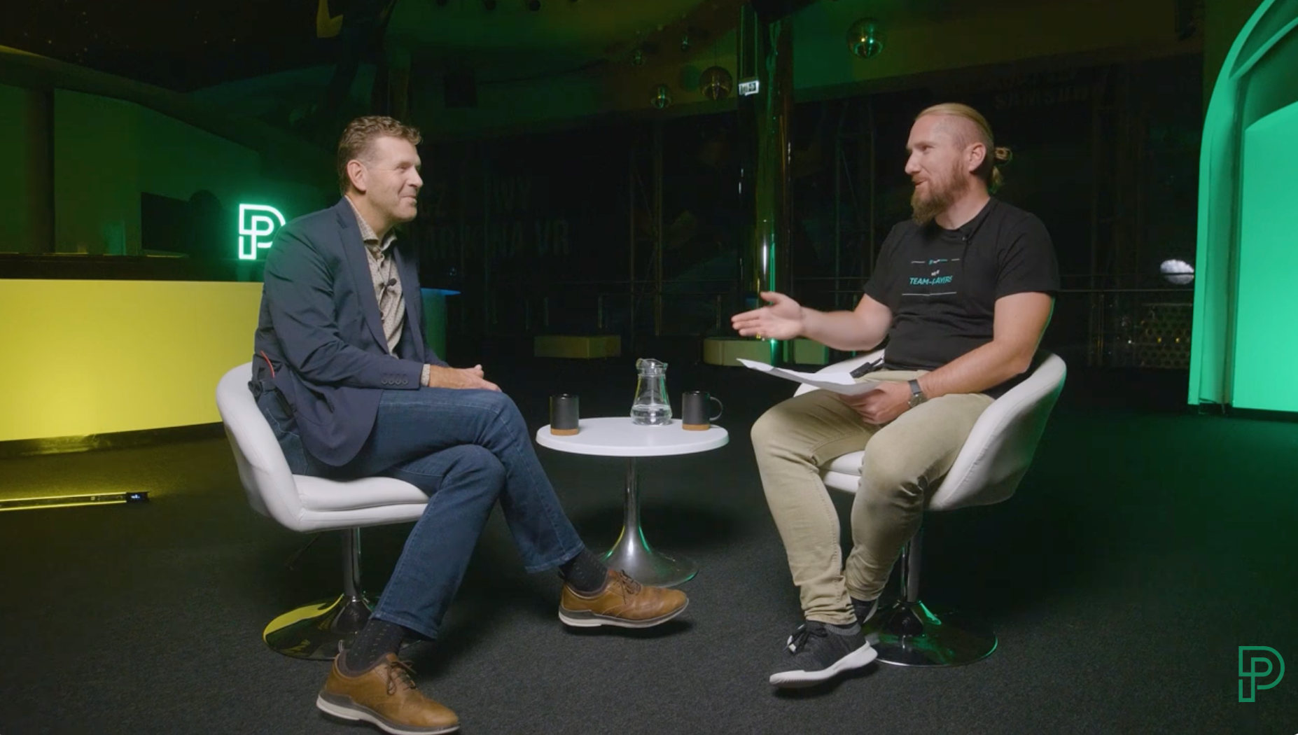 Two people sit in white chairs, facing each other to talk in a studio with green lighting and a round table between them.