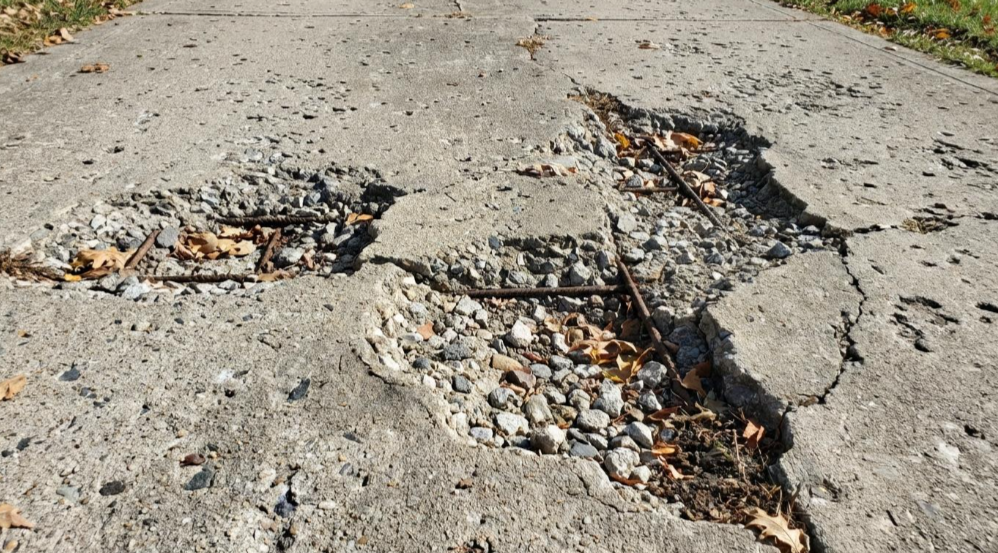Cracked concrete road with potholes, exposed gravel, and broken asphalt patches