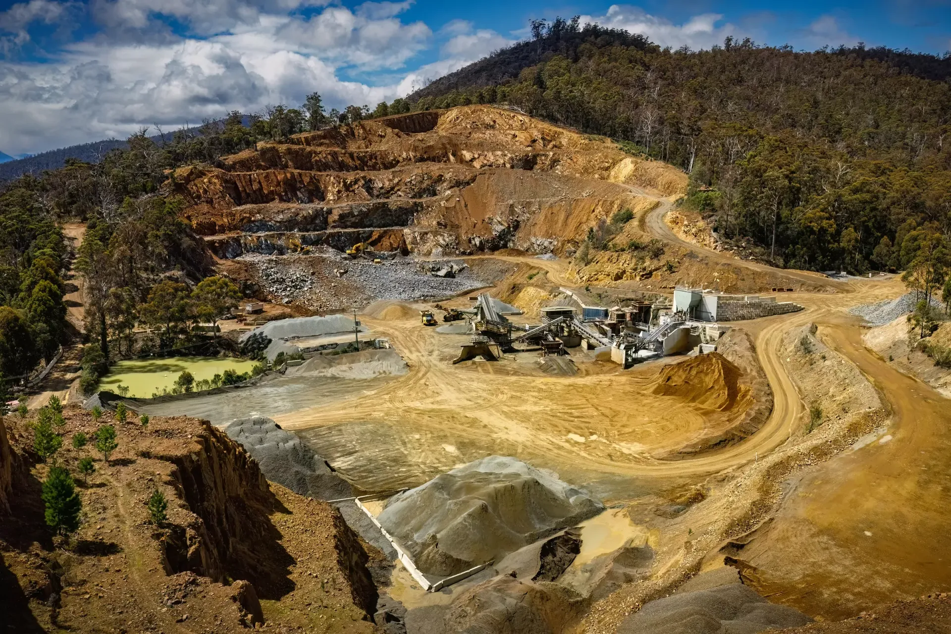 Terraced open-pit mine with excavated earth and machinery beneath a wooded hillside and blue sky