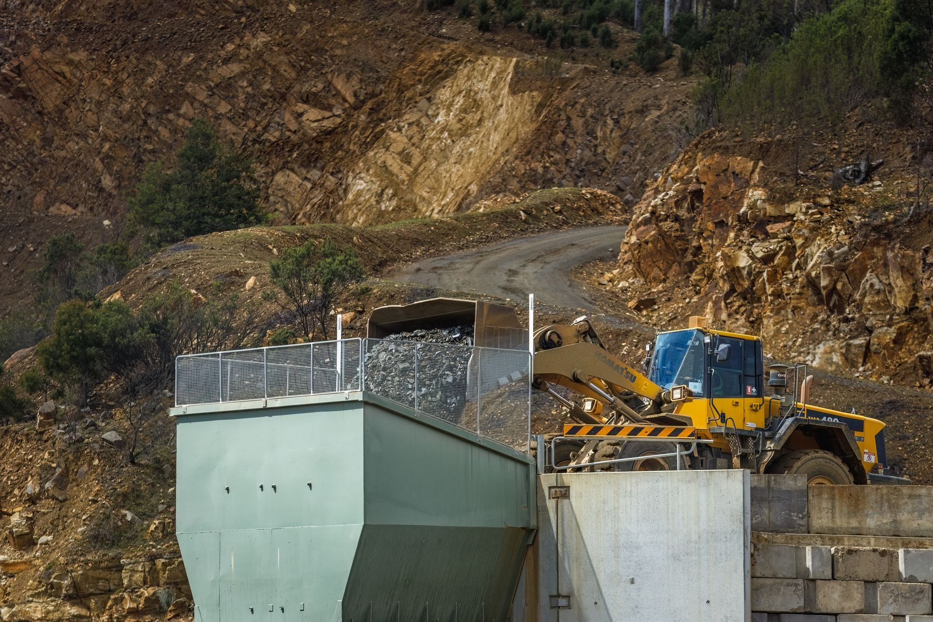 Yellow excavator on a rocky construction site beside a concrete retaining wall, with hillside behind.