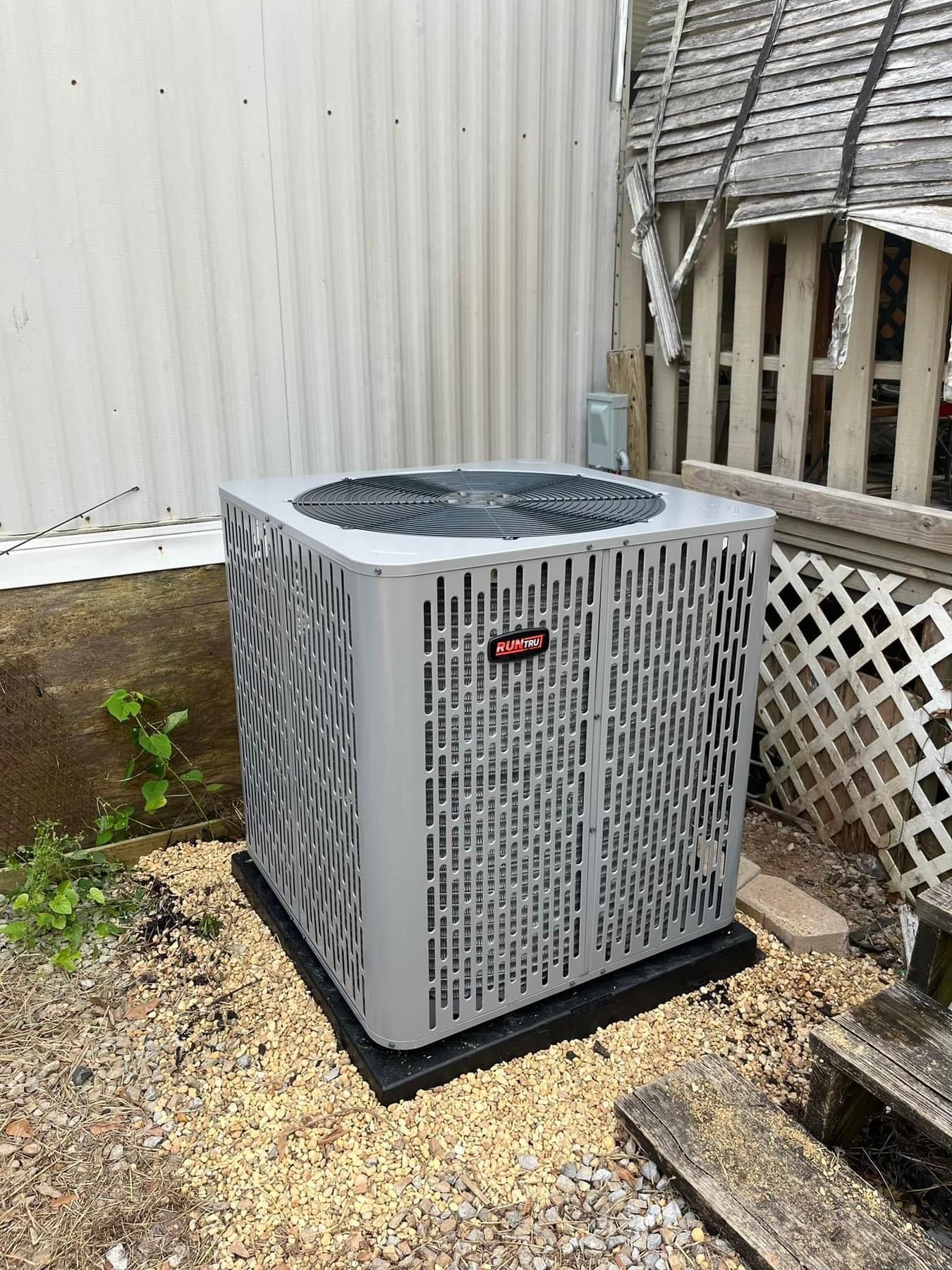 A large air conditioner is sitting on top of a pile of gravel in a backyard.