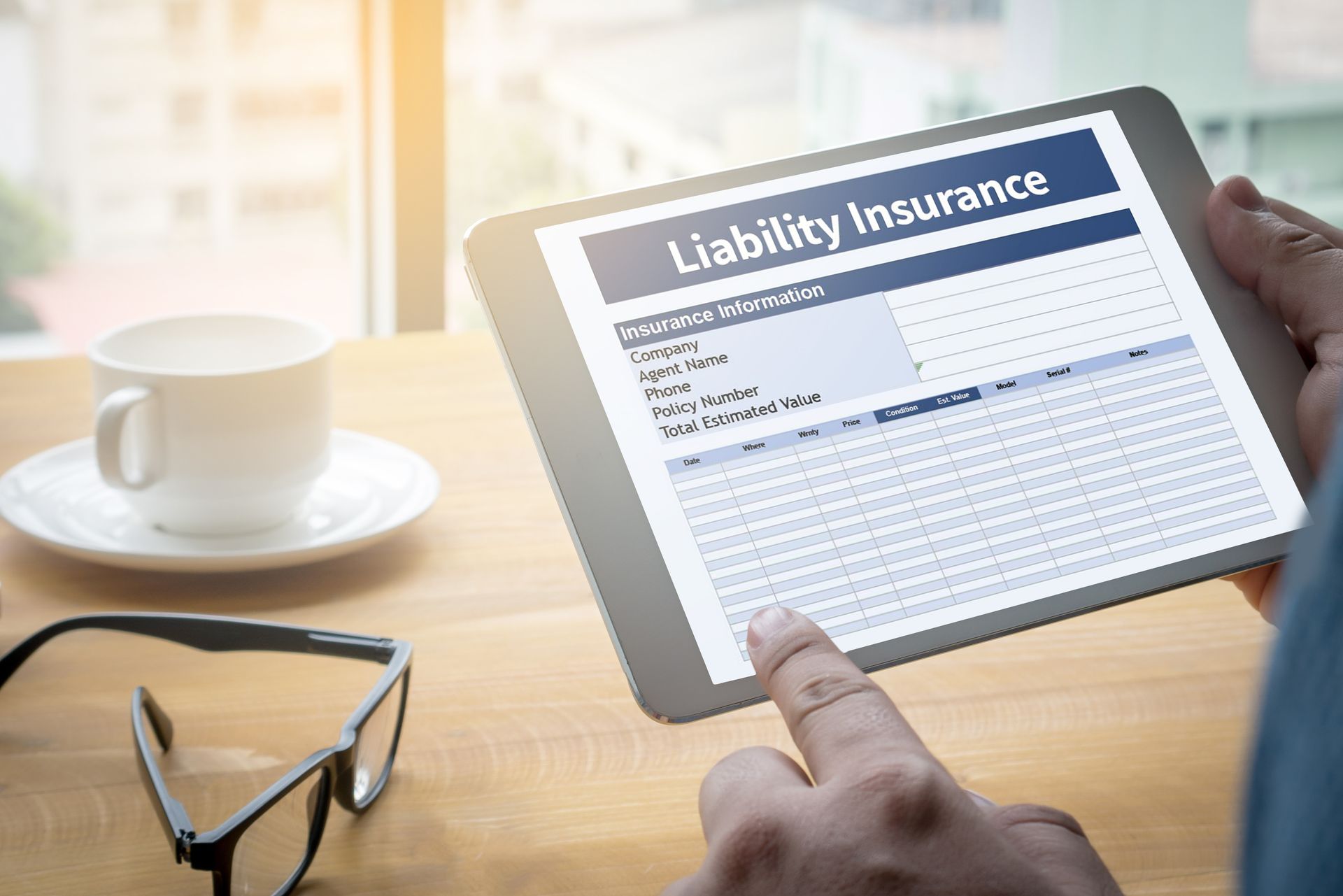 A person holds a tablet displaying a liability insurance form. A cup of coffee and glasses sit nearby on a desk.