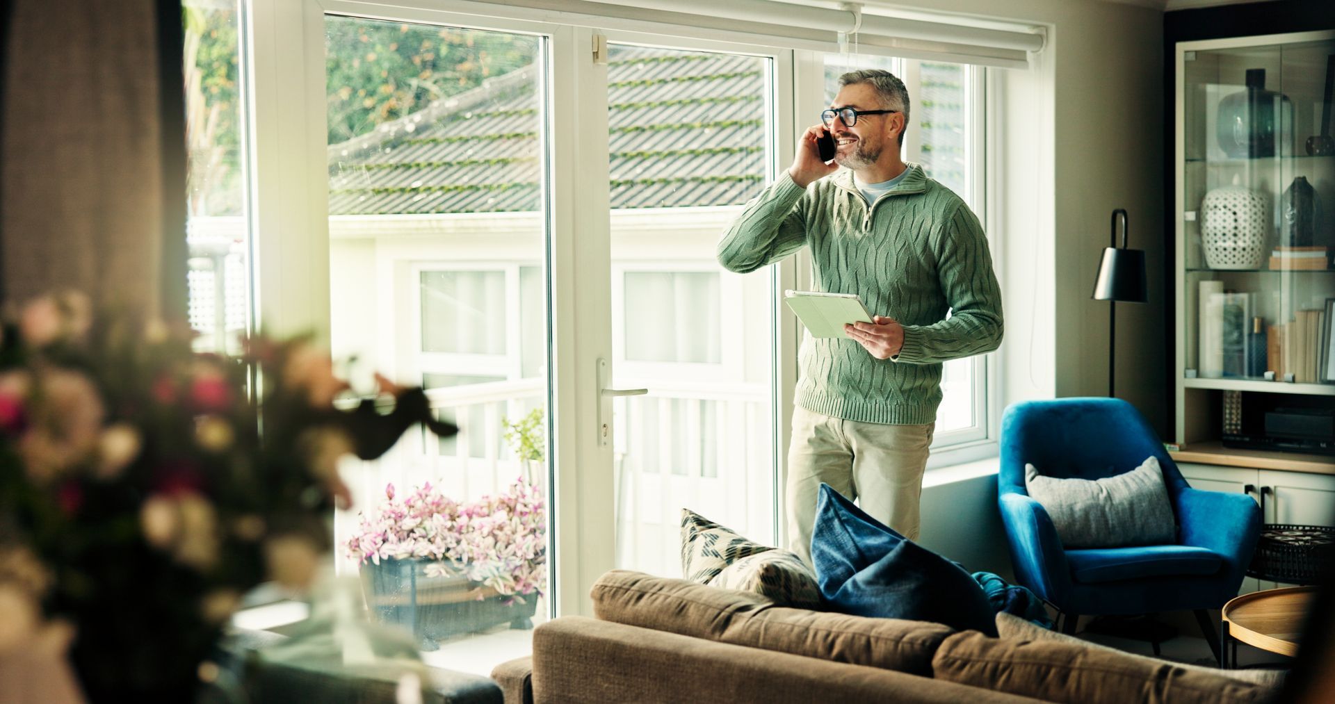 Man talking on phone by window, holding papers. Inside a home, sweater, glasses.