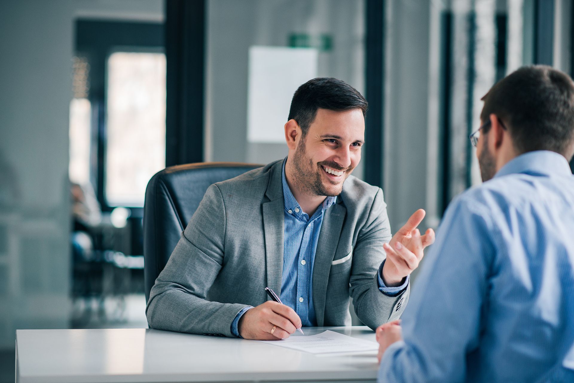 Man in gray blazer smiling, talking with another person across a desk. Office setting.