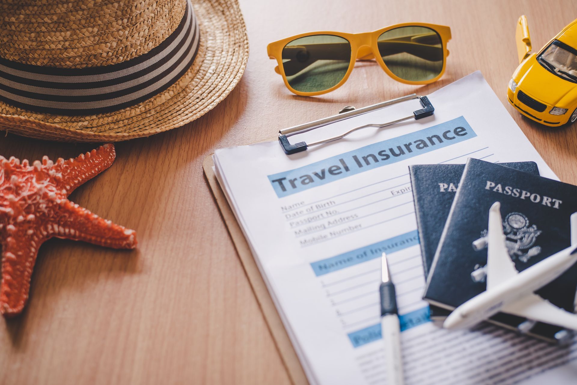 Travel insurance paperwork, passports, sunglasses, toy airplane, and hat on wooden surface.