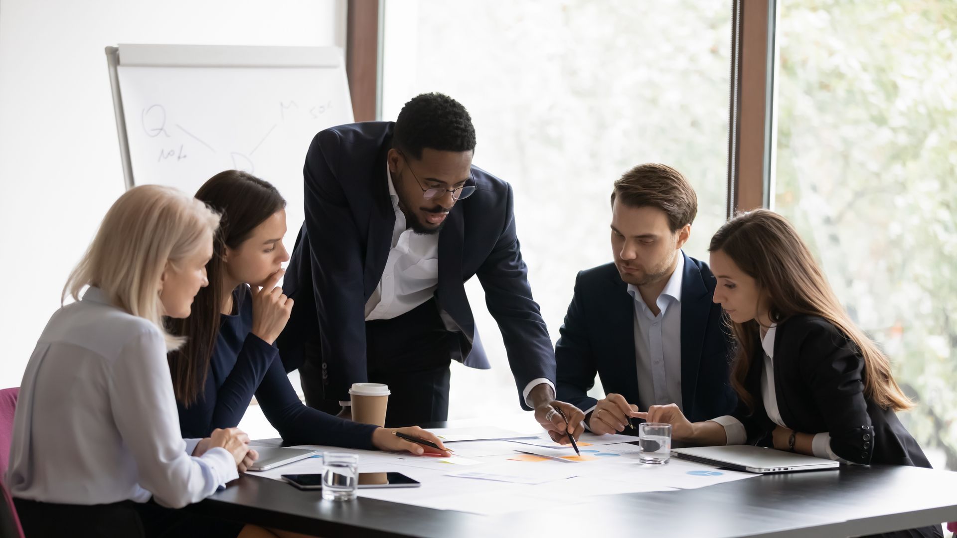Business team reviewing documents at a table. A man gestures at paperwork, others look on intently.