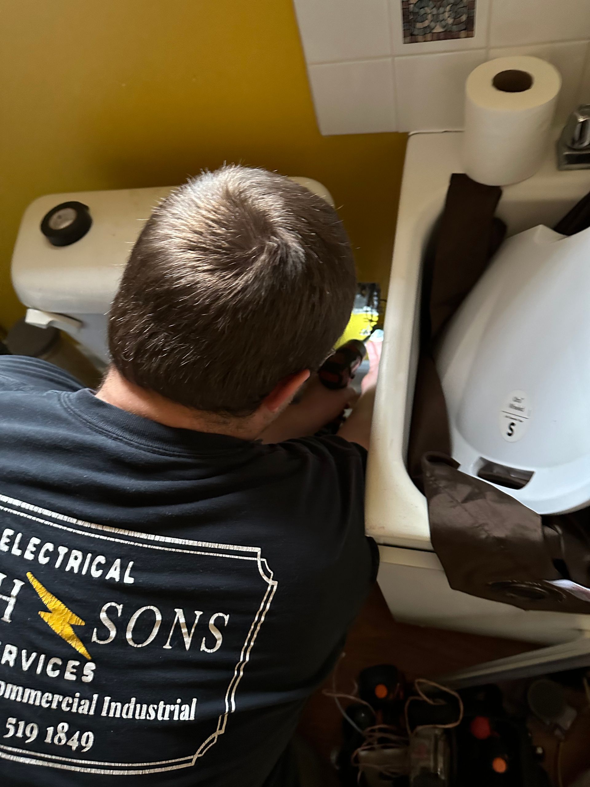 Licensed electrician troubleshooting wiring behind wall near restroom sink and toilet