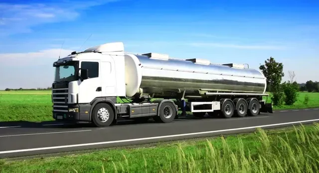 Autocisterna bianca su strada, che trasporta liquidi; campi verdi e cielo azzurro sullo sfondo.
