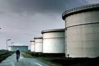 Un uomo in bicicletta passa accanto a grandi serbatoi industriali sotto un cielo nuvoloso.