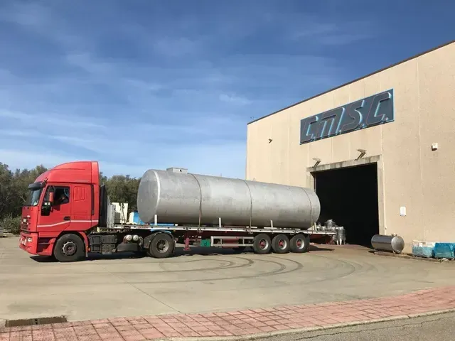 Un camion rosso che trasporta un grande carro armato argentato, parcheggiato fuori da un edificio chiaro.
