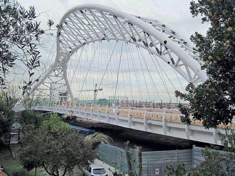 Ponte ad arco bianco con cavi di sospensione. Cielo grigio, alberi su entrambi i lati, lavori in corso sullo sfondo.