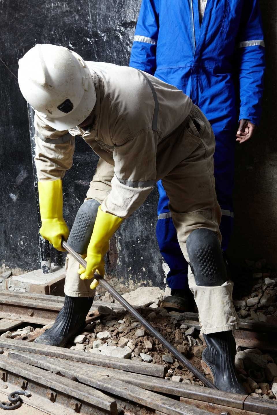 Un lavoratore con equipaggiamento protettivo utilizza una barra di metallo, con un altro lavoratore sullo sfondo.