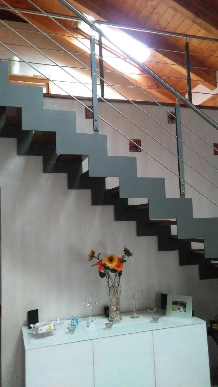 Scala sospesa grigia e nera con ringhiera in metallo, che sale verso un soffitto in legno.