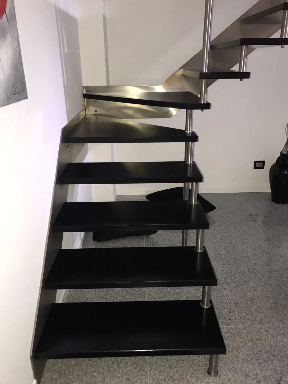 Scala in legno nero con supporti in acciaio inossidabile contro una parete bianca e un pavimento grigio.