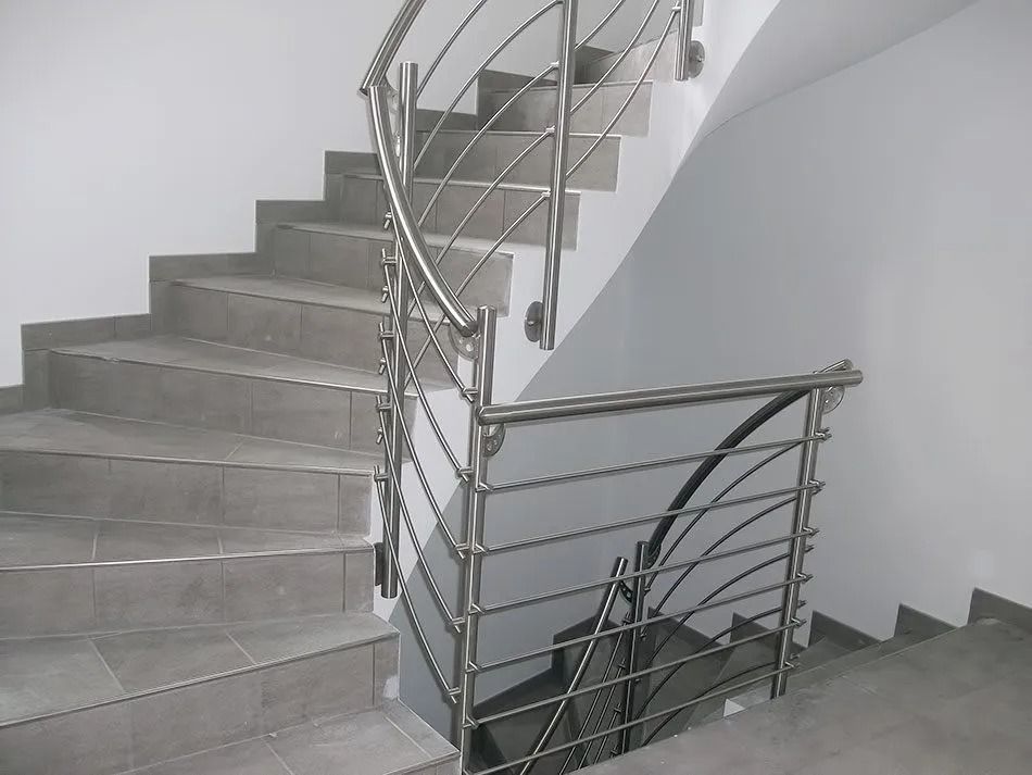 Scala a chiocciola rivestita di piastrelle grigie con corrimano in acciaio inossidabile contro pareti bianche.