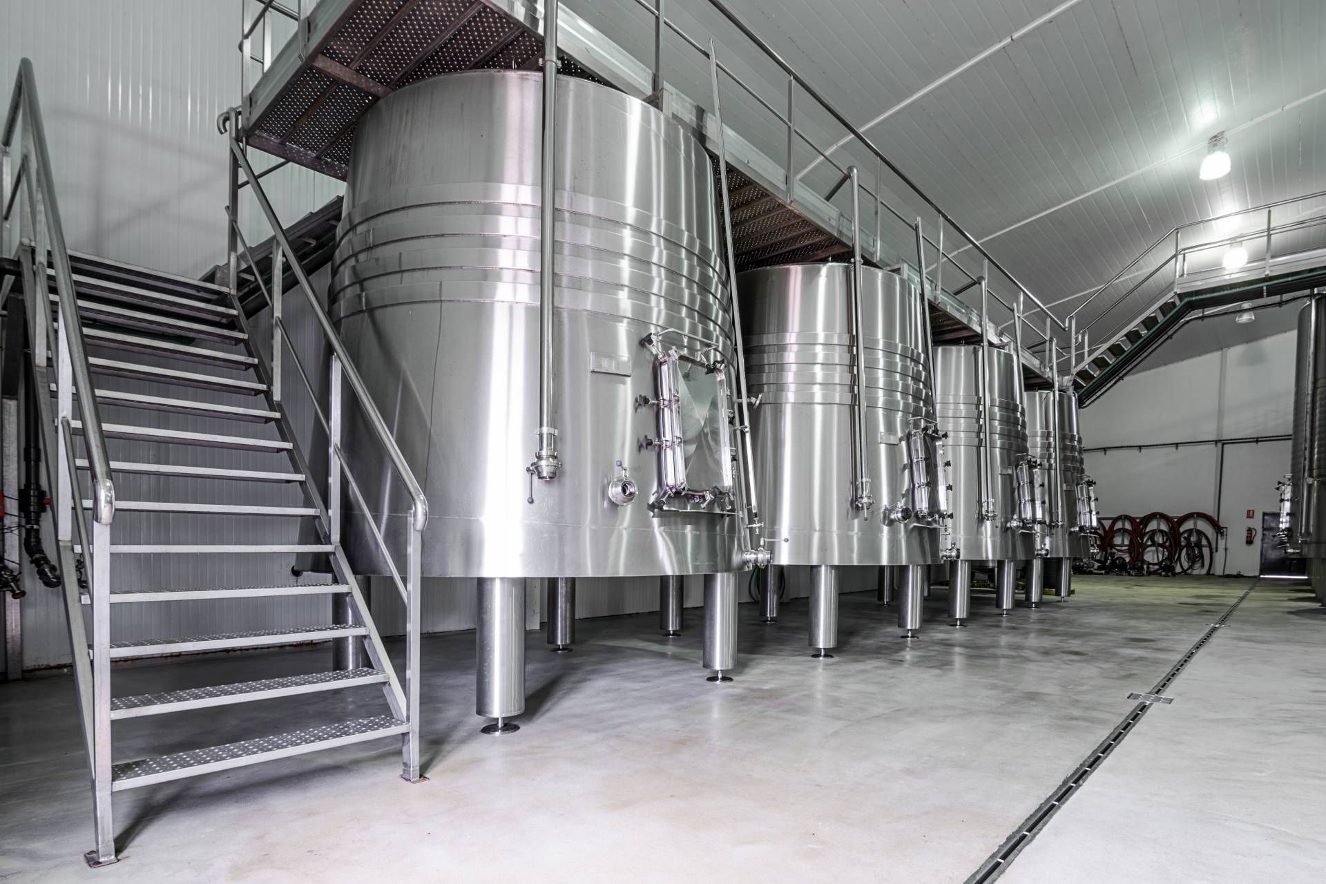 Fila di serbatoi di fermentazione in acciaio inossidabile in un magazzino, con un pallet di legno accatastato sullo sfondo.