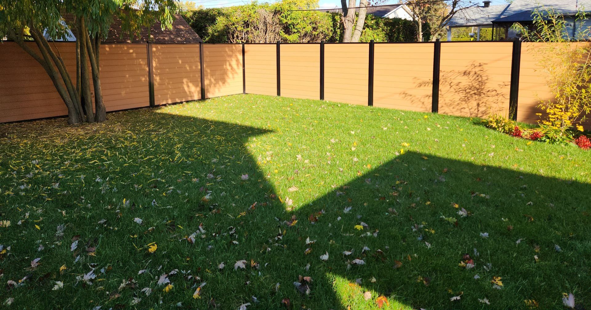 Clôture en bois entourant une pelouse verte recouverte de feuilles mortes, avec un arbre et du soleil.