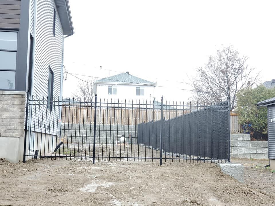 Une clôture noire entoure une cour en terre devant une maison.