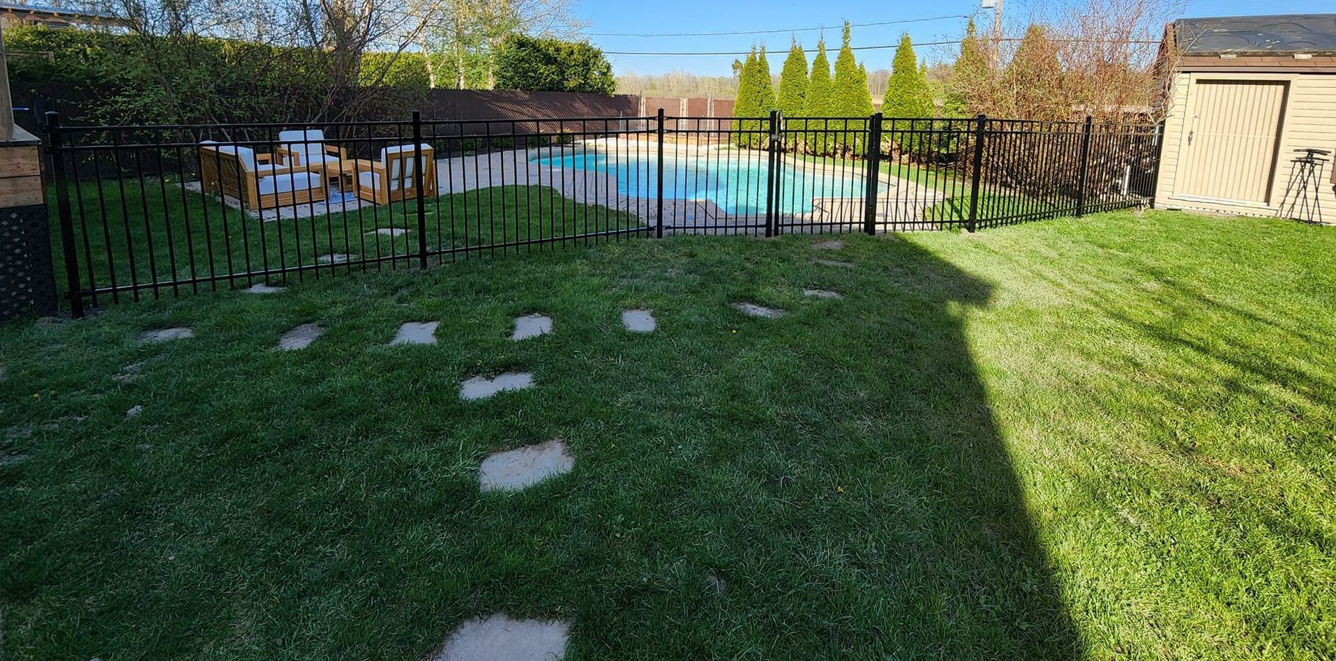 Une cour arrière avec piscine, clôture noire, pas japonais sur la pelouse et abri de jardin, par une journée ensoleillée.