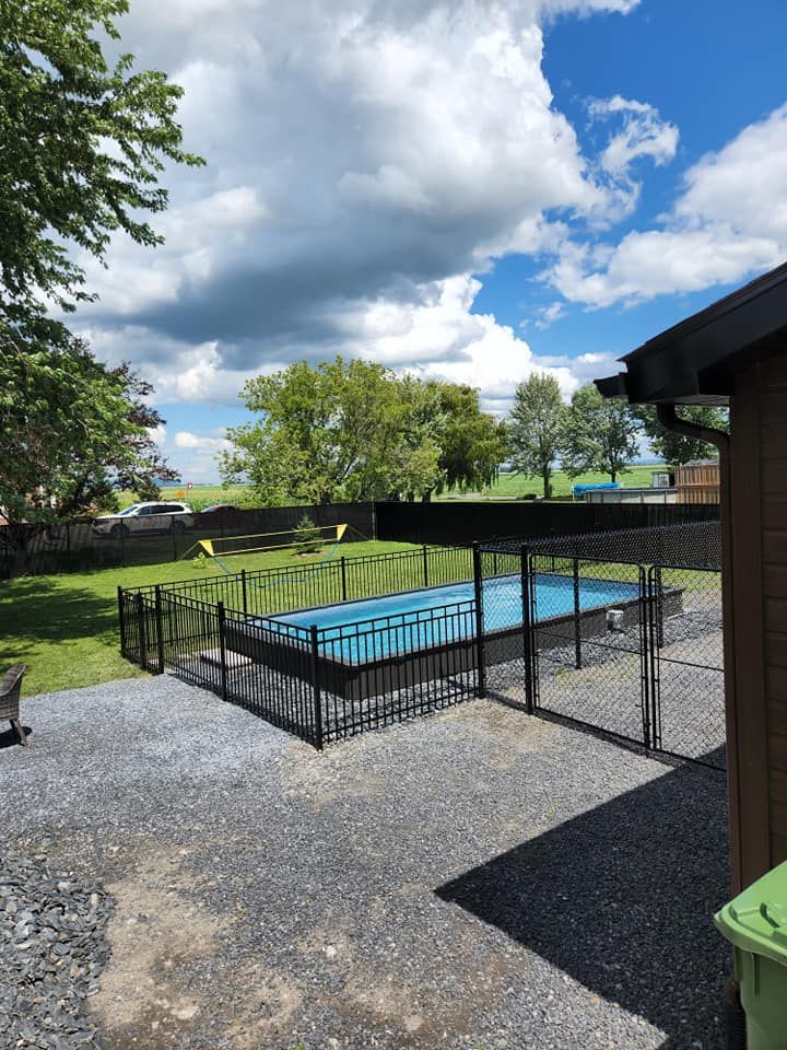 Piscine entourée d'une clôture noire sur une terrasse en gravier. Pelouse verte, arbres et ciel bleu nuageux en arrière-plan.