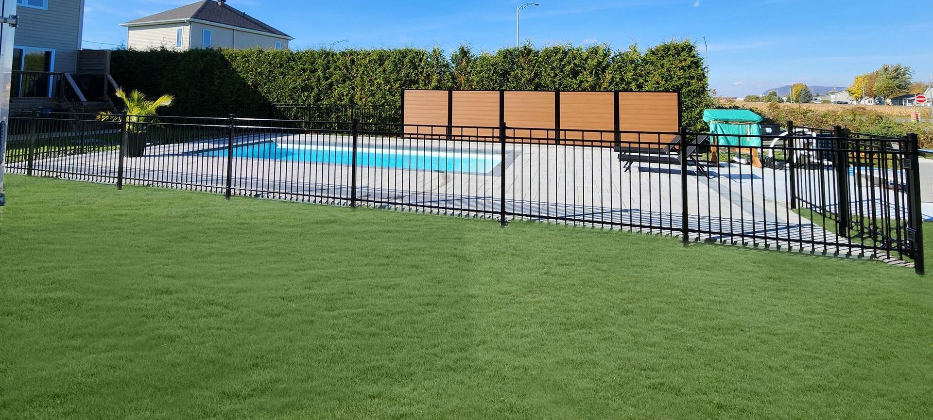 Jardin avec piscine, clôture noire, pelouse verte, arbres et ciel bleu.