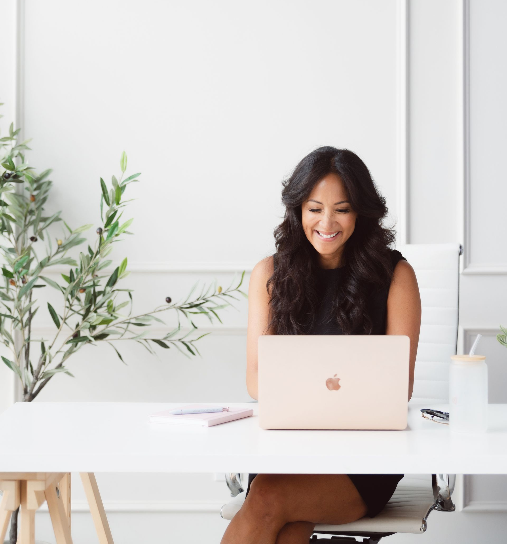 Woman smiling at a laptop, sitting at a white desk in a bright office; olive tree in the background.