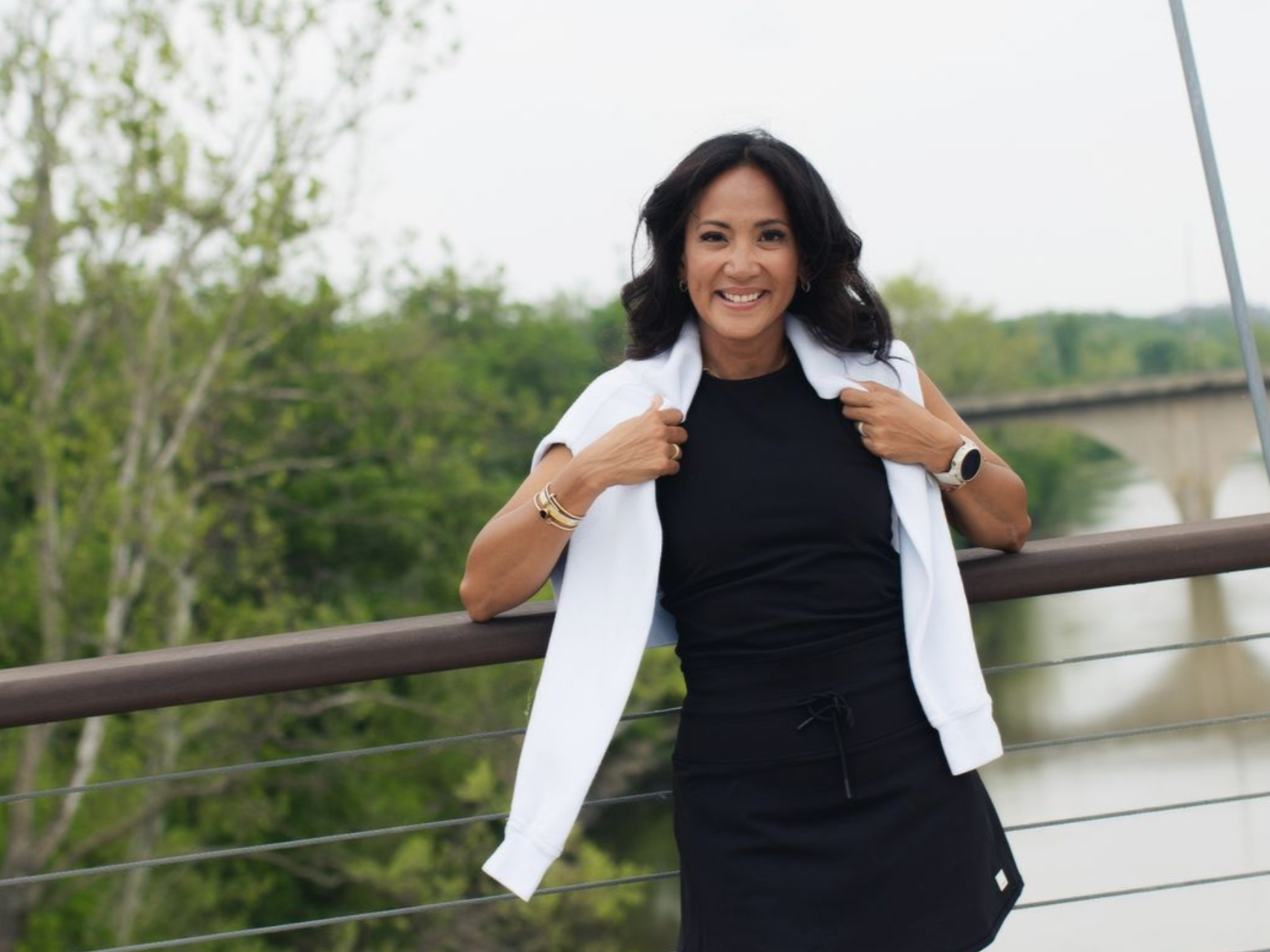 Woman smiling, wearing black dress, white jacket, outdoors on bridge.