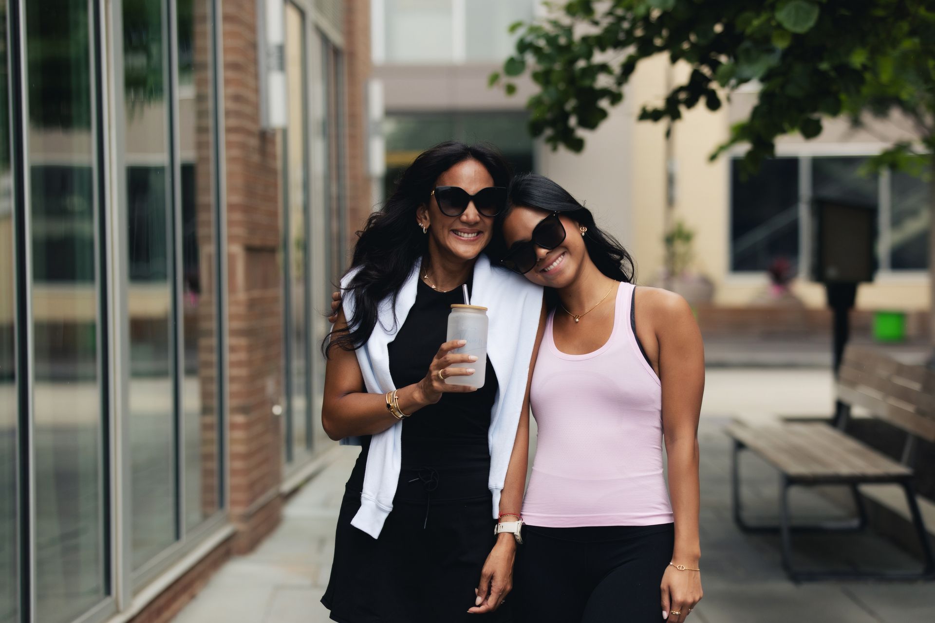 Two women smiling, walking outside. One holds a beverage. Brick building backdrop.