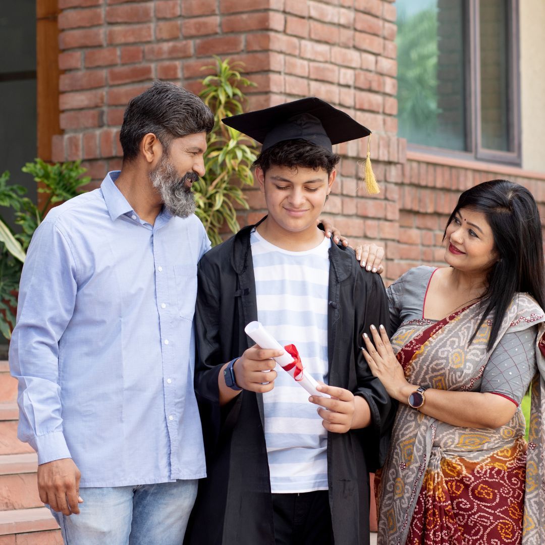 Family celebrating graduation. Son in cap and gown holds diploma, parents smile, embrace him outdoors.