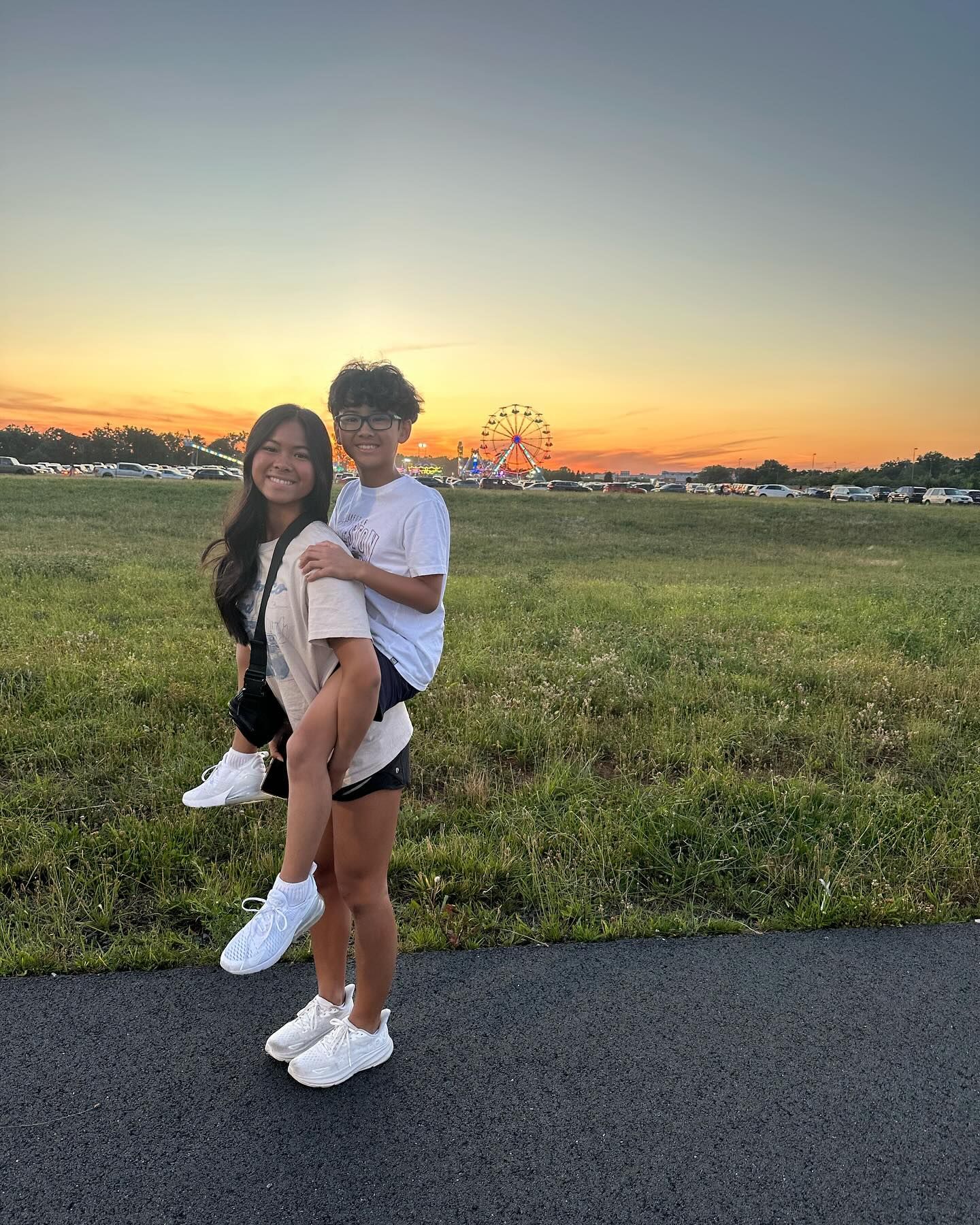 Woman giving piggyback ride to person with glasses, sunset and Ferris wheel in background.
