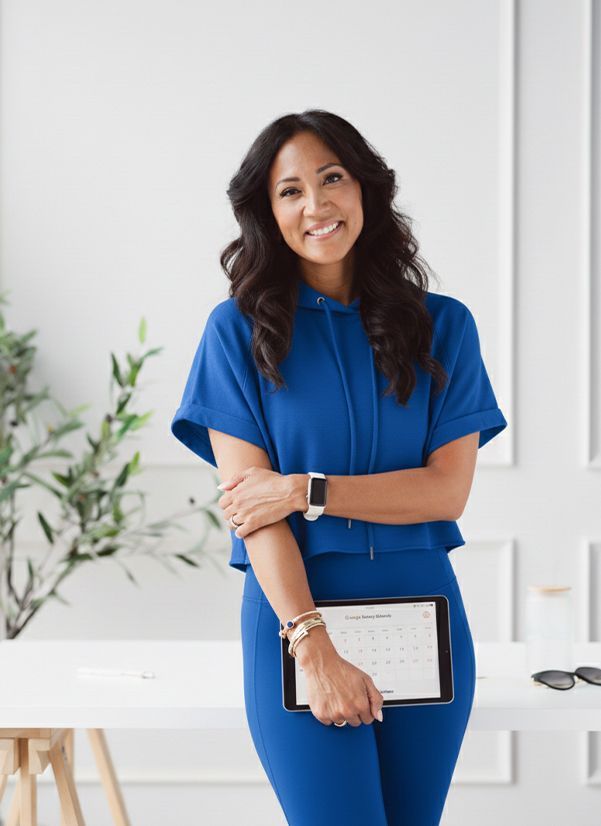 Woman in blue outfit smiling, holding tablet with calendar, standing in a bright office space.