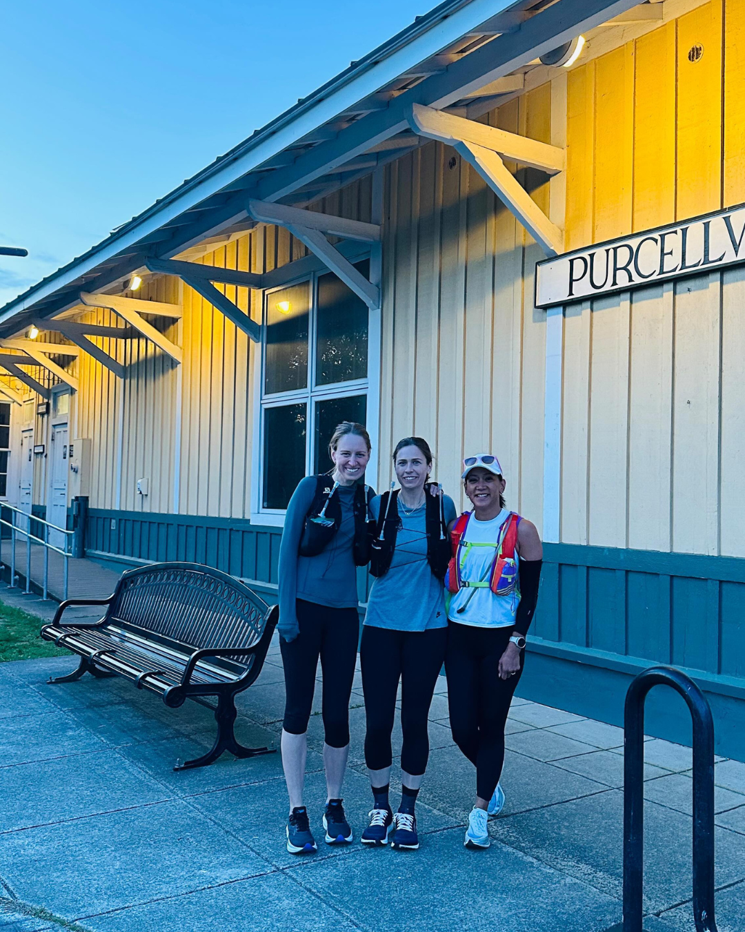 Three runners pose in front of a Purcellville station. They wear running gear.