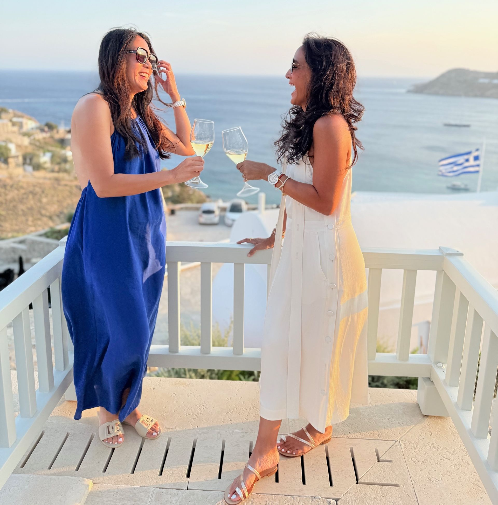 Two people on a balcony, toasting with wine, ocean view. One in blue dress, the other in white, sunny.