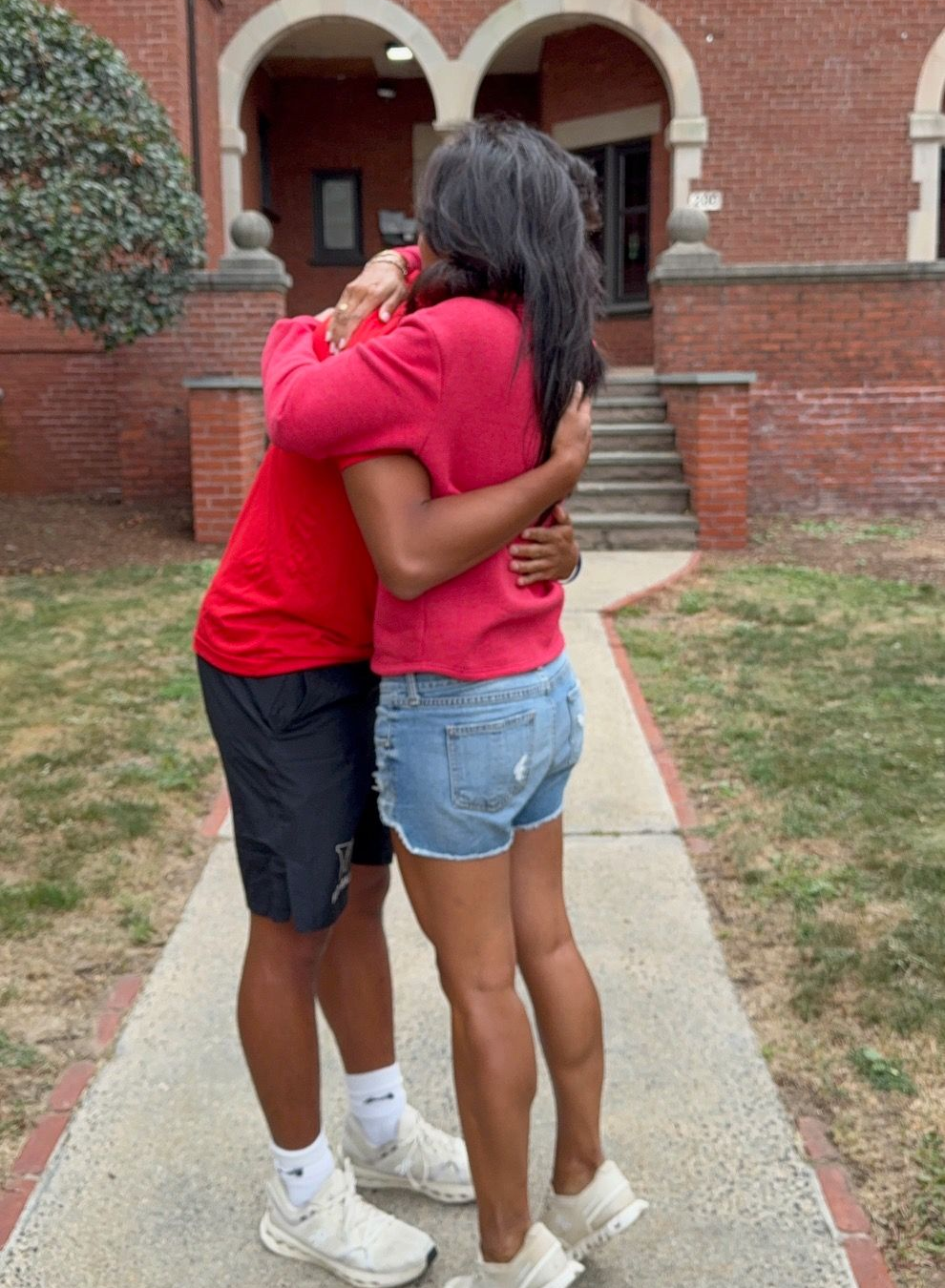 Two people embracing on a walkway in front of a brick building. One wears red, the other, denim shorts.