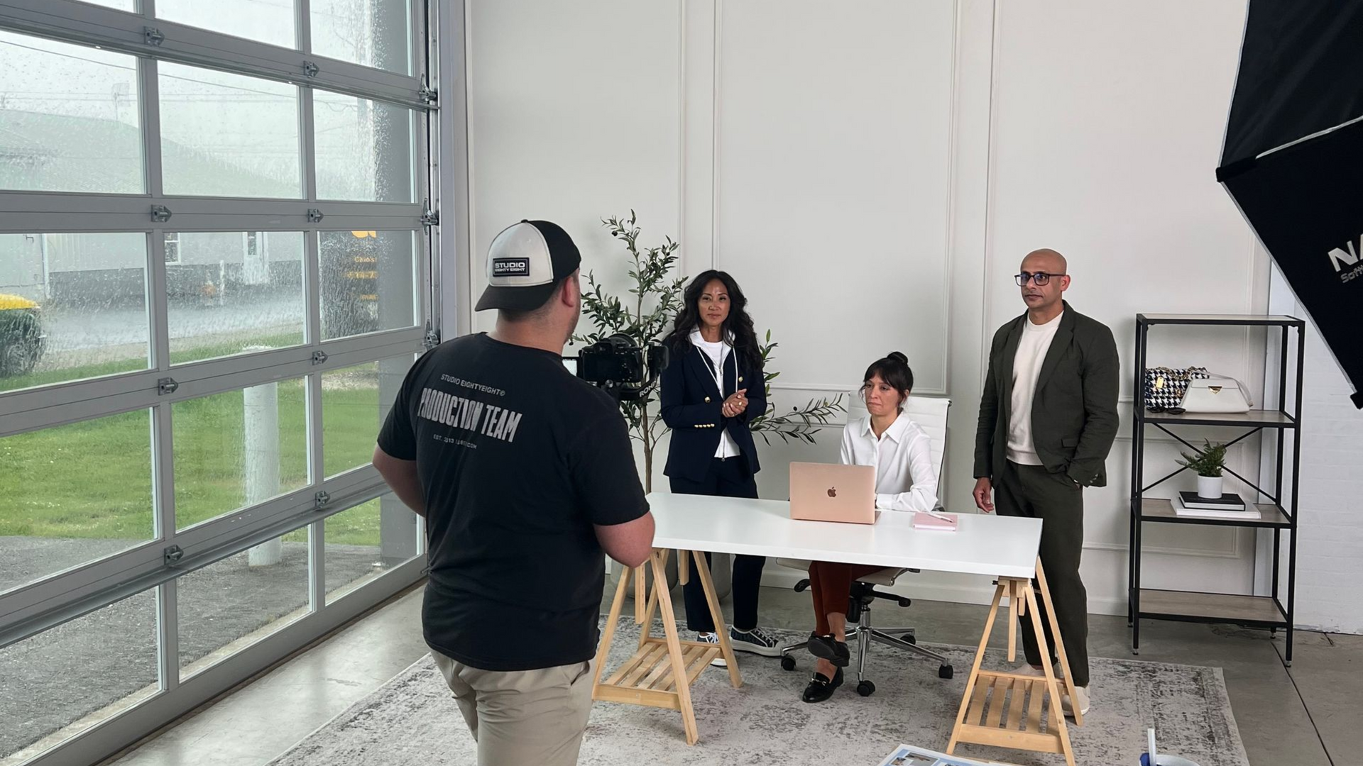 People posing for a photo shoot in a studio. One person at a desk, two standing, and one filming. White walls, gray floor.