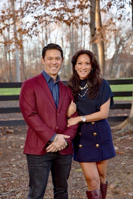 Person in dark glasses reads braille, with a person assisting, in a library with bookshelves.