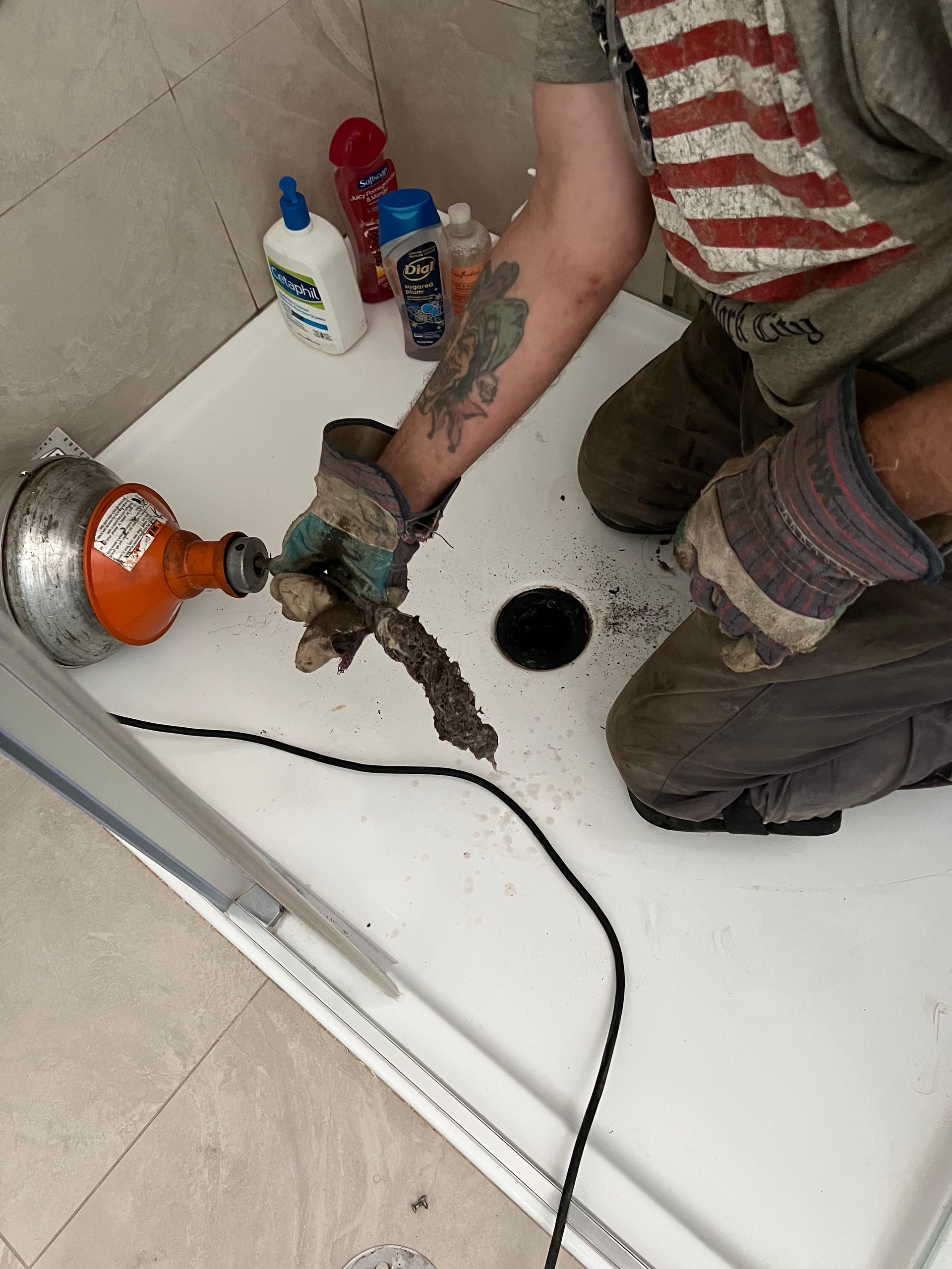 Plumber unclogs shower drain with a snake. Person in work gloves and an American flag shirt.
