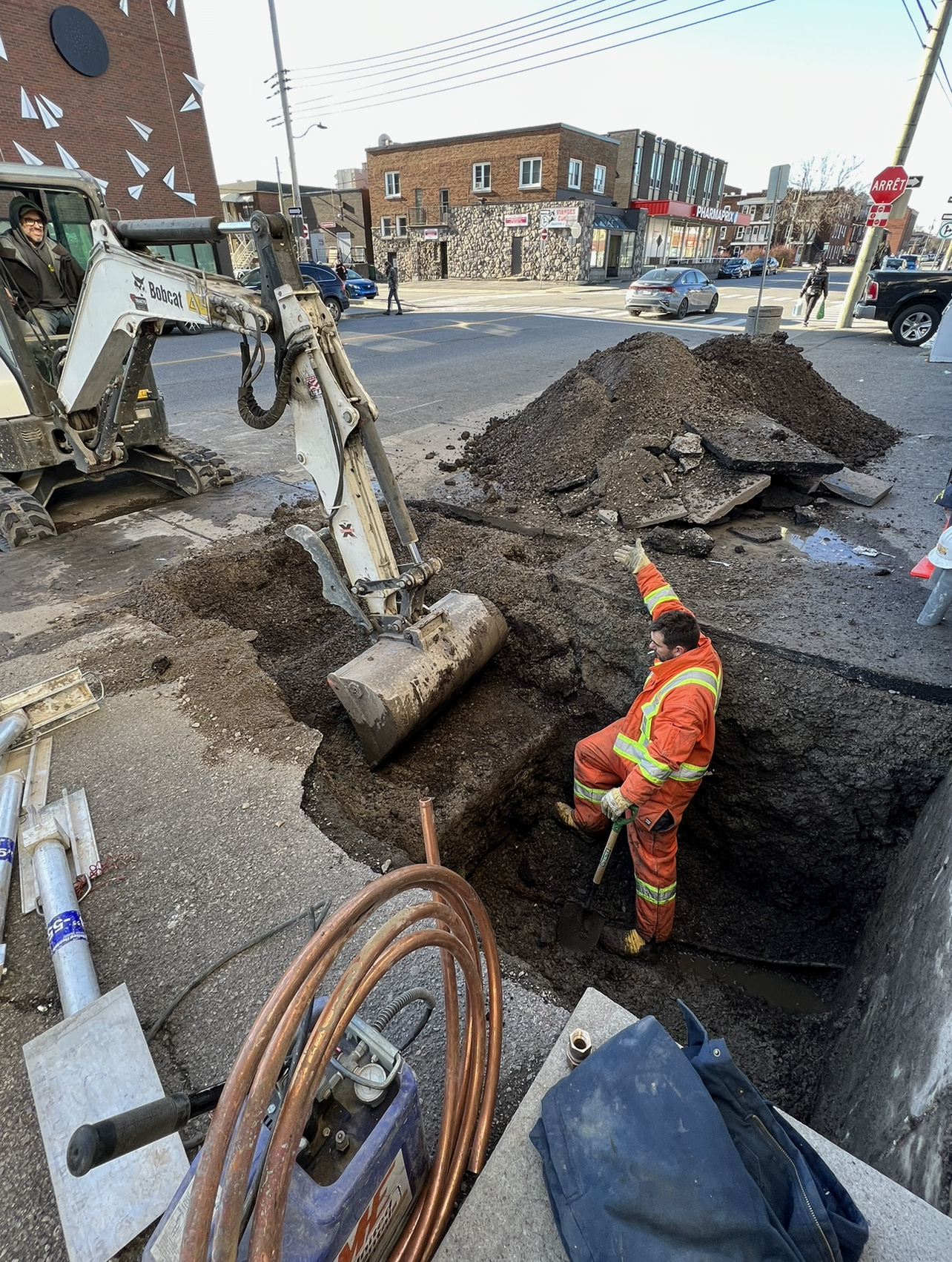 Man in orange suit working in a deep construction pit, excavator nearby on street.