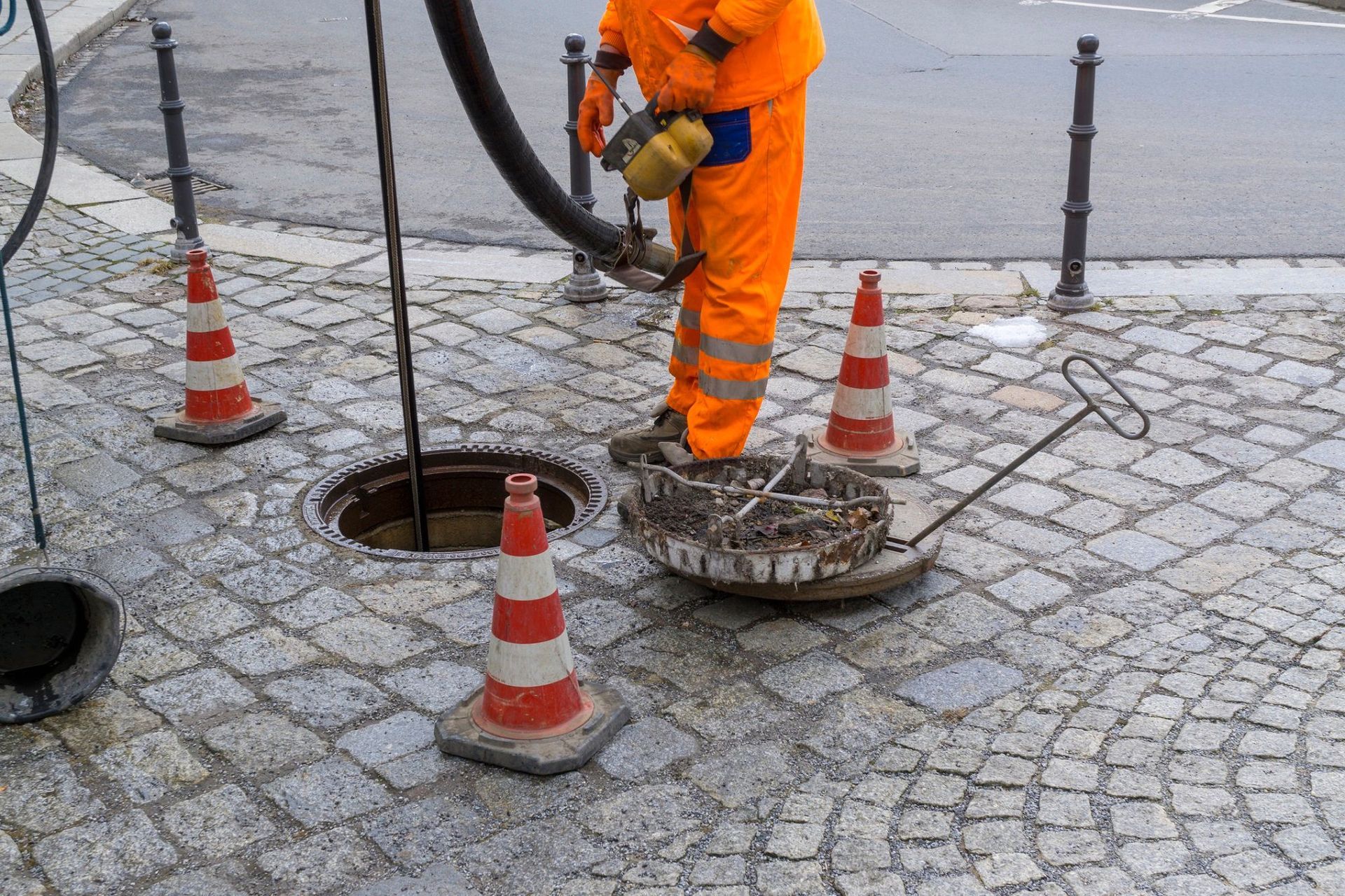 Worker in orange suit working on a manhole in cobblestone street, using vacuum hose, traffic cones present.