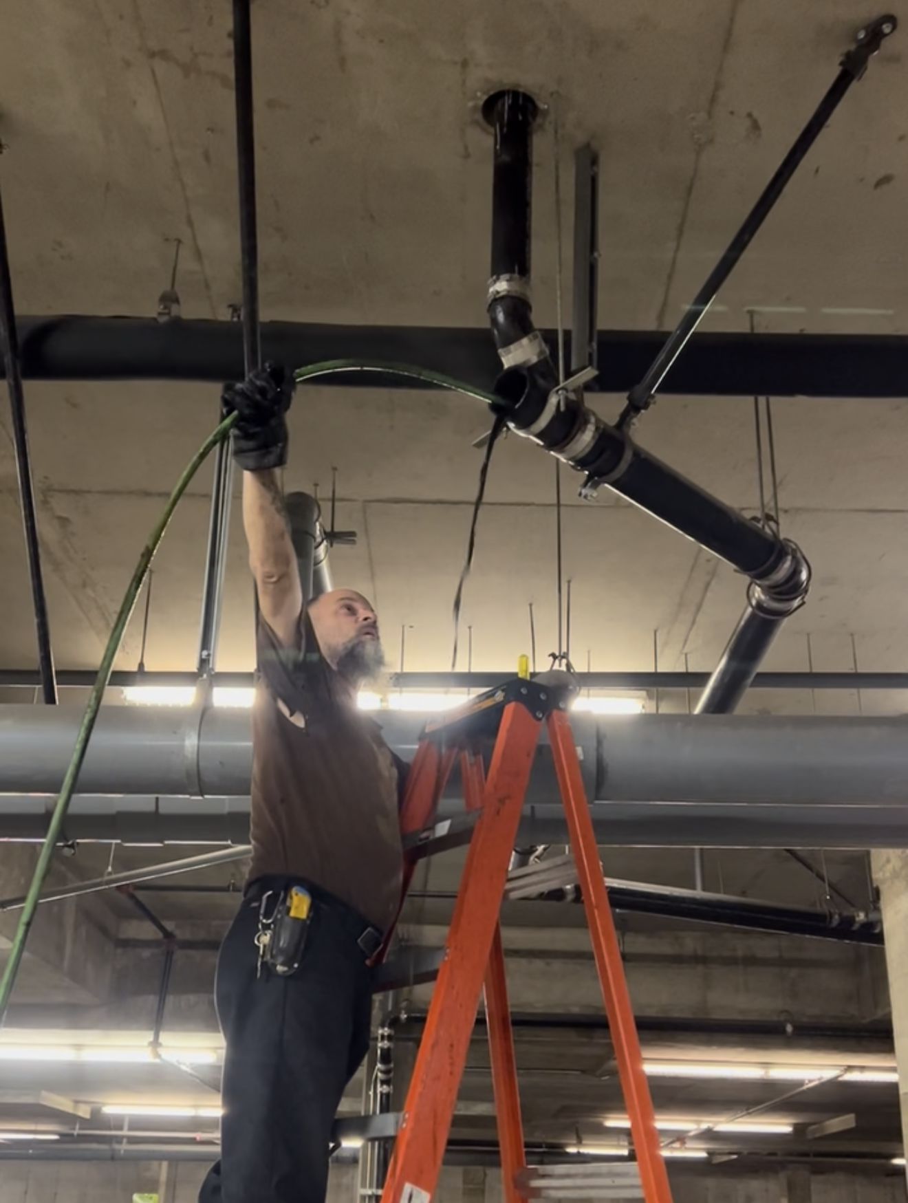 Construction worker on a ladder reaching up to work on black pipes near a concrete ceiling.