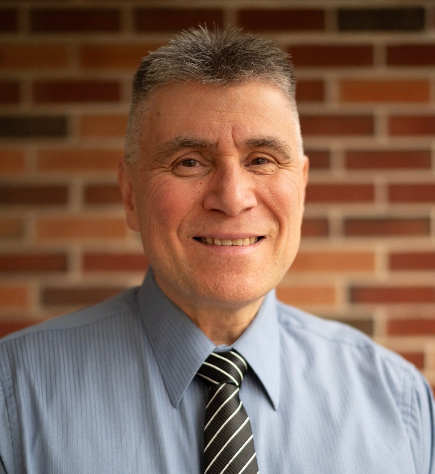 A man in a blue shirt and tie is smiling in front of a brick wall.