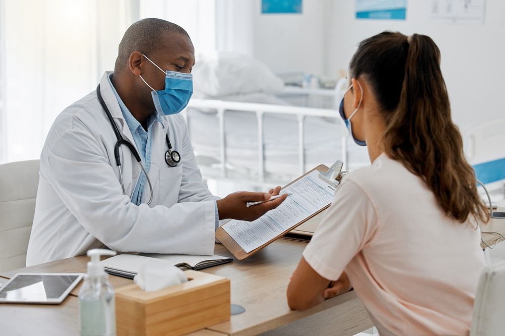 Doctor in mask reviewing paperwork with a patient at a desk in a medical office.