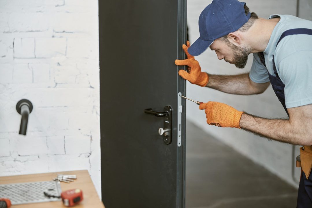 Man in blue cap and gloves installing a door lock with a screwdriver.