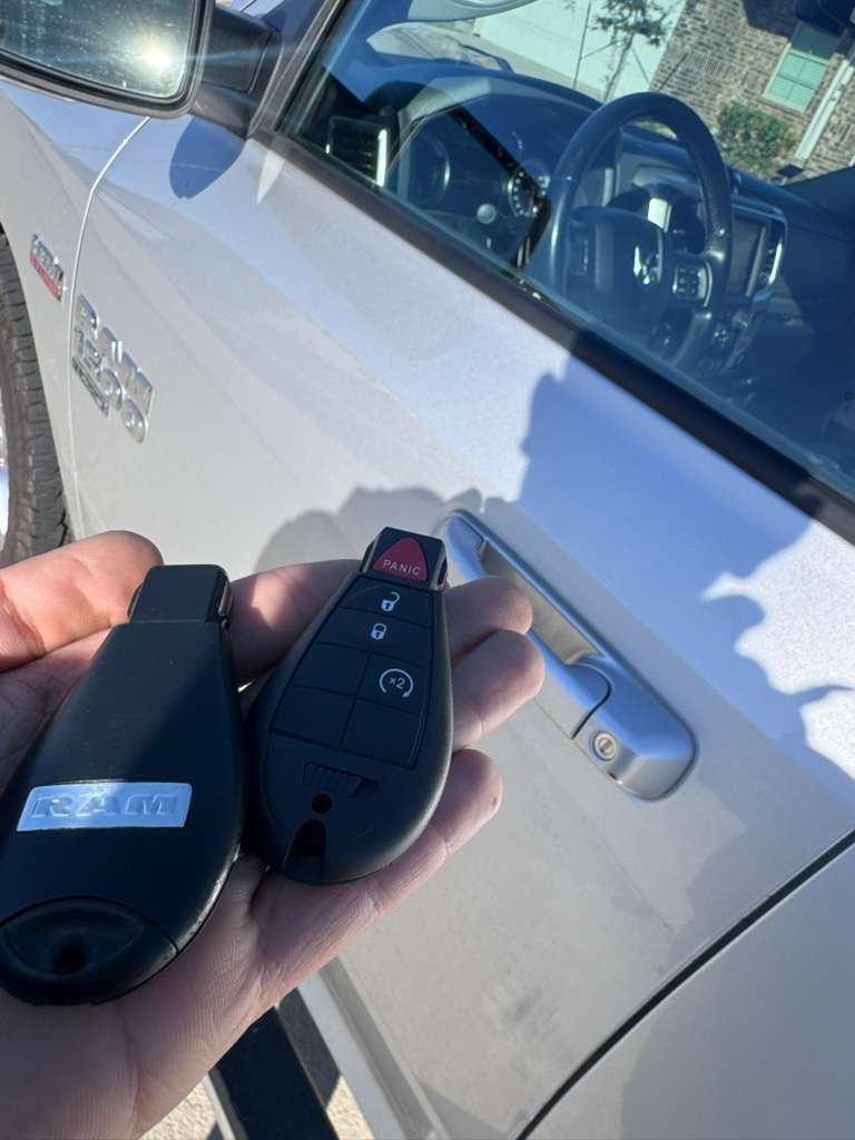 Hand holding two car keys in front of a silver Ram truck. One key is black, the other has buttons.