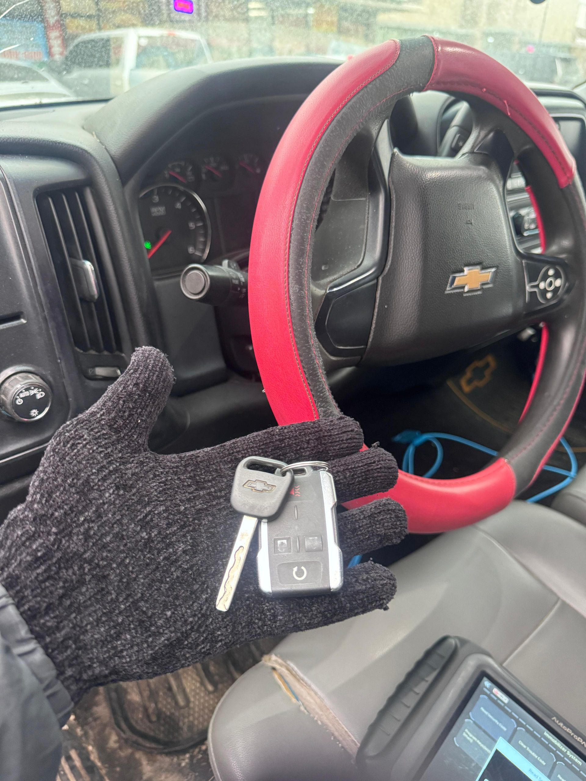 Gloved hand holding car keys inside a Chevy Silverado. Steering wheel with red cover visible, diagnostic tool in view.