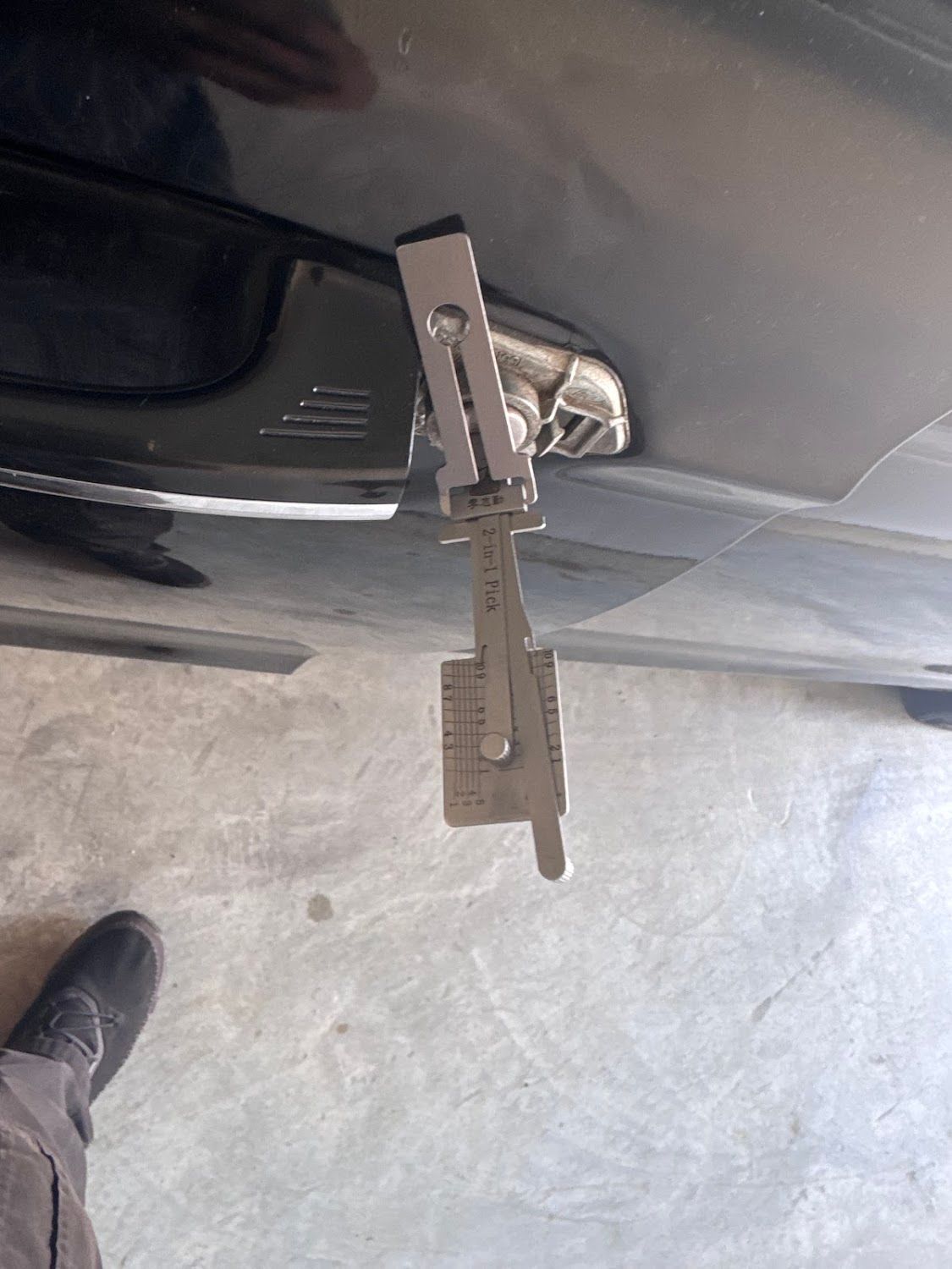 A lock pick tool inserted into a car door lock, black car, concrete ground, black shoe visible.